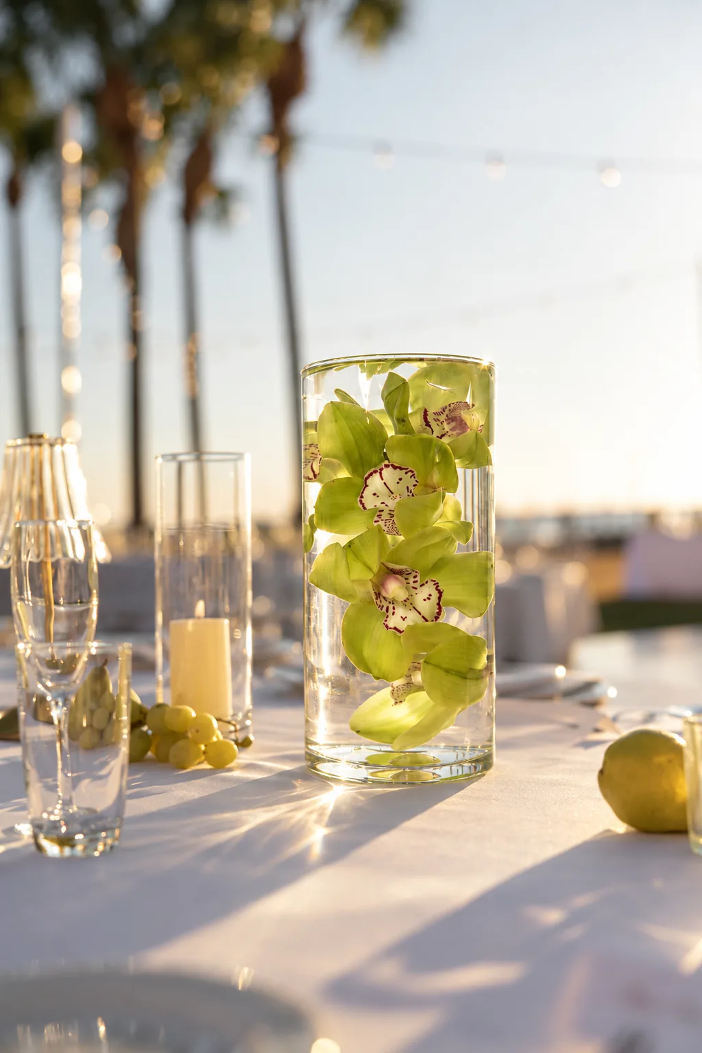 Composition with pale green orchids submerged in a water-filled glass cylinder on a set table outdoors