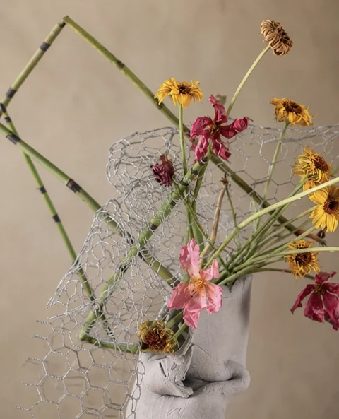 Floral study with crossed green stems, wire mesh and orange and pink flowers in a white vase