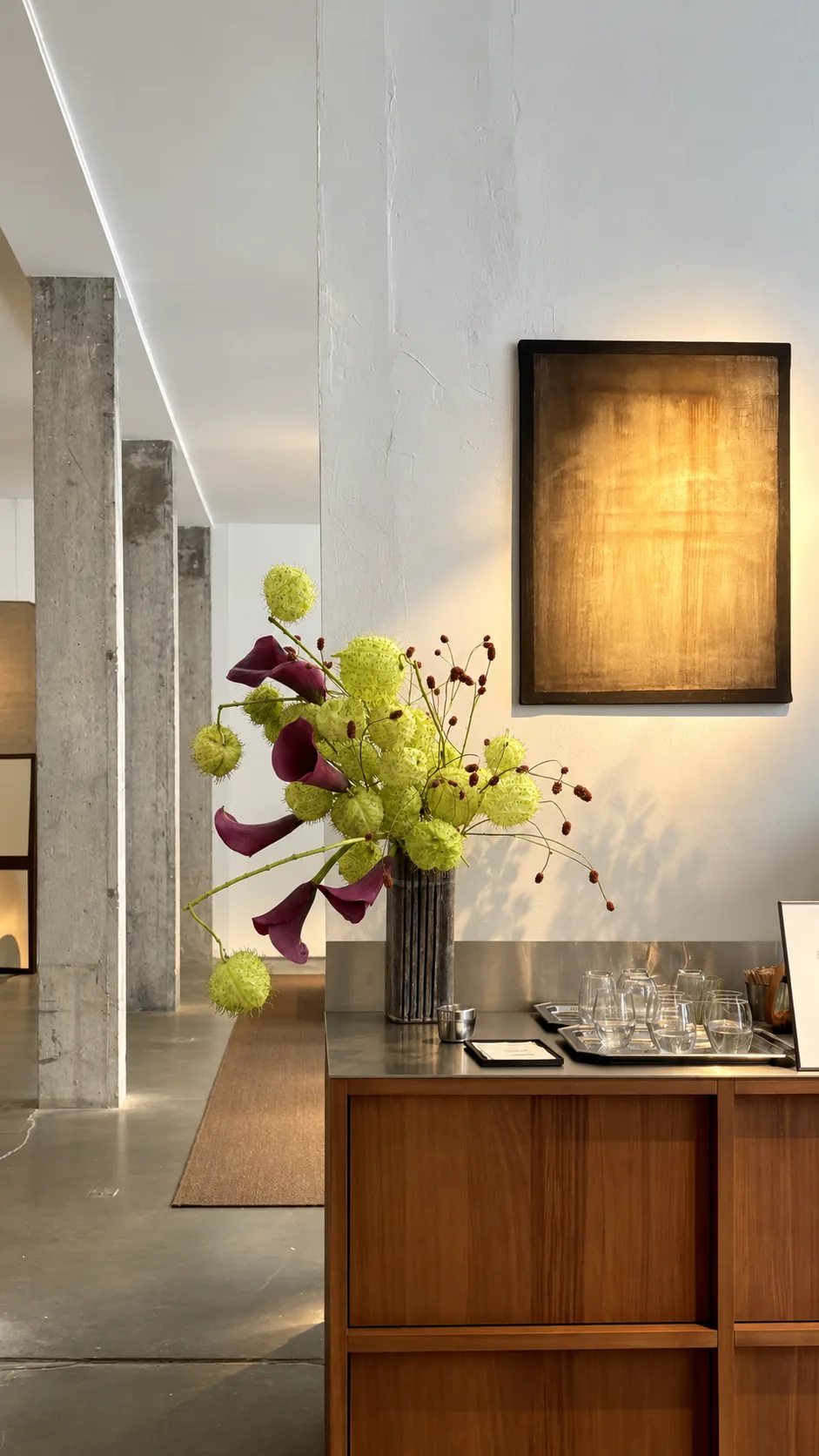 Composition with magenta callas and spiky green seed pods in a dark vase on a wooden sideboard in an exhibition space with concrete columns