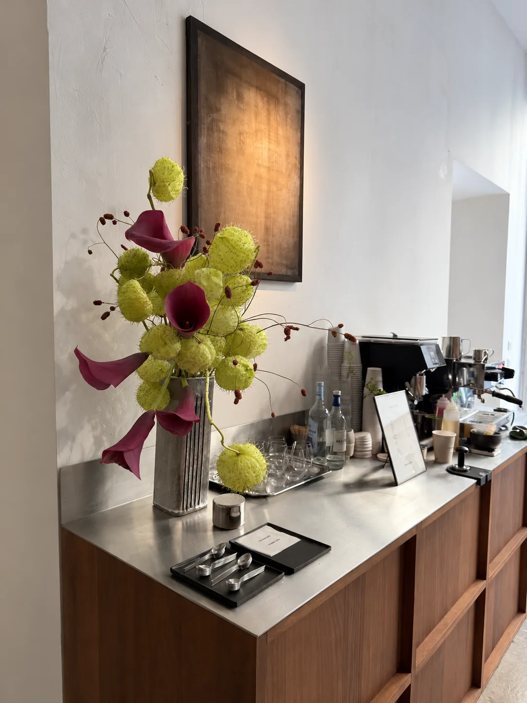 Composition with magenta callas, spiky green seed pods and red branches in a silvered vase on a steel counter in an interior space